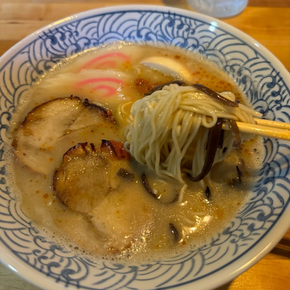 Chopsticks lifting ramen noodles from a bowl with pork, fish cake, and broth.