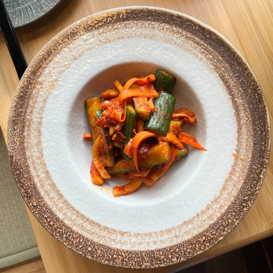 A white bowl with a brown rim holds a vibrant mix of kimchi-style cucumbers and carrots.