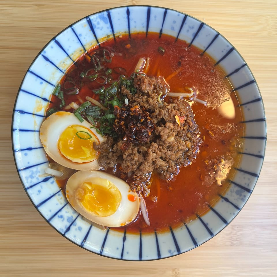 A bowl of Ramen Thukpa ramen with a rich red broth, ground meat, green onions, and two soft-boiled eggs.